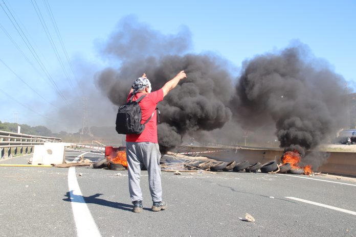 Un trabajador del metal ante una barricada de neumáticos ardiendo durante la primera jornada de huelga indefinida en el Valle de Escombreras, en Cartagena. | Foto: Dominic David Skerrett. Un trabajador del metal ante una barricada de neumáticos ardiendo durante la primera jornada de huelga indefinida en el Valle de Escombreras, en Cartagena. | Foto: Dominic David Skerrett.